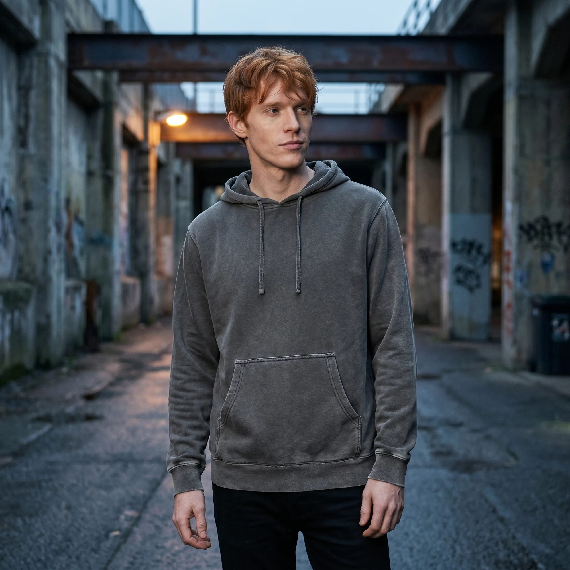 Man wearing a black pigment dye hoodie, front view, hood down, in a moody industrial setting