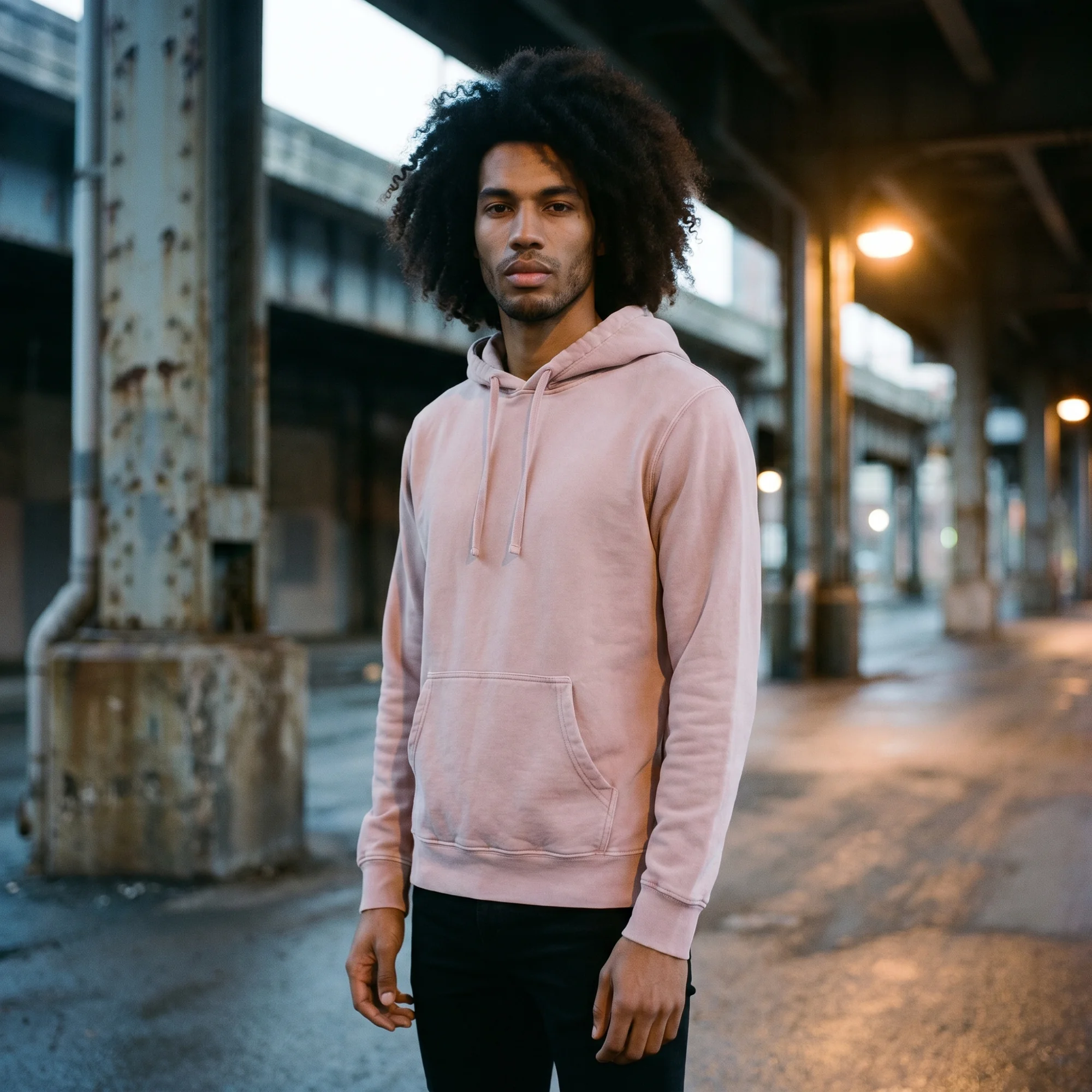 Man wearing a pink pigment dye hoodie, front view, hood down, in a moody industrial setting