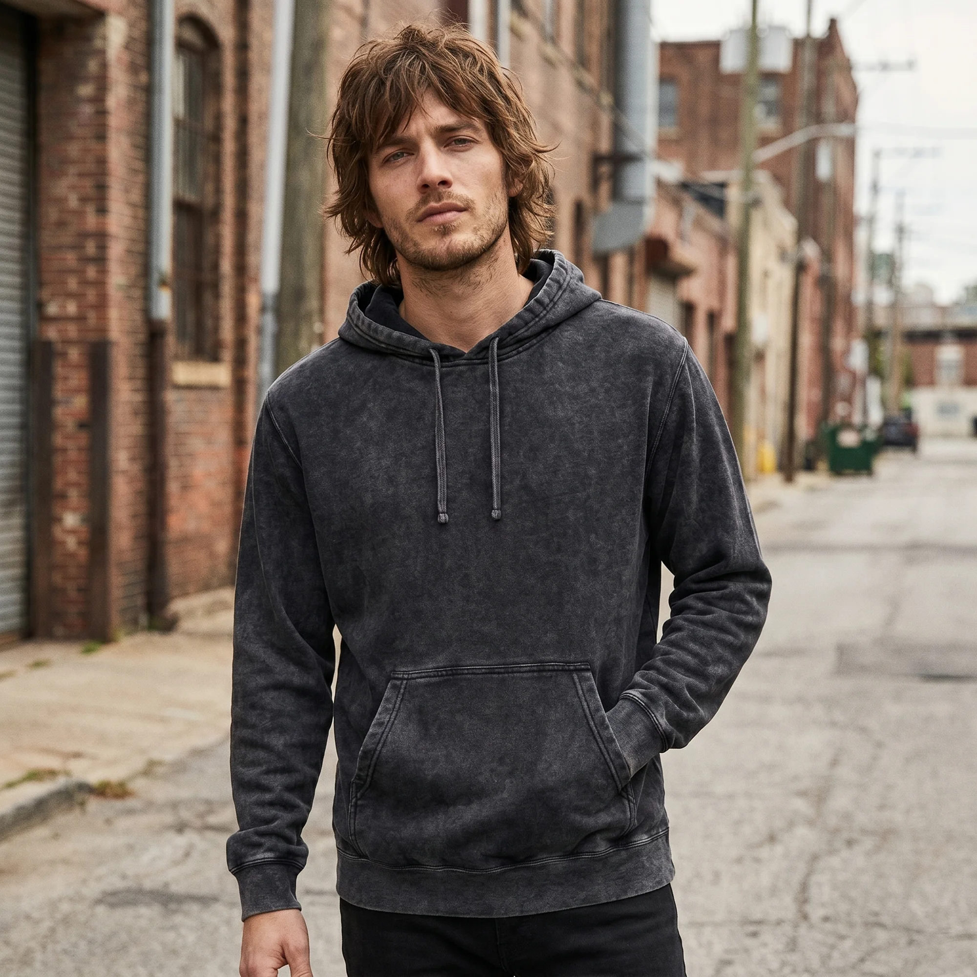 Man wearing a black mineral wash hoodie, front view, hood down, walking down a brick alley