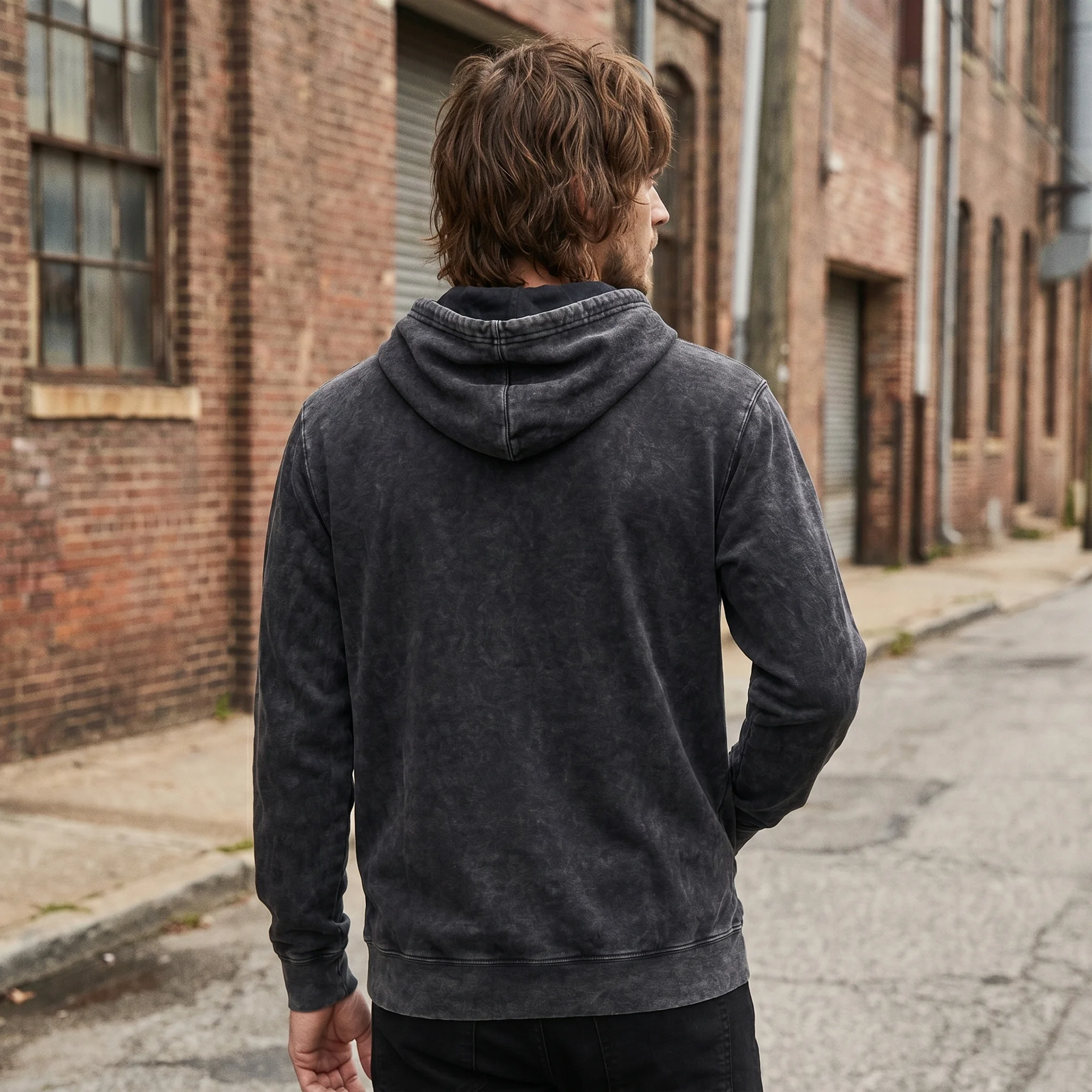 Back view of a man wearing a black mineral wash hoodie with the hood down, in a brick alley