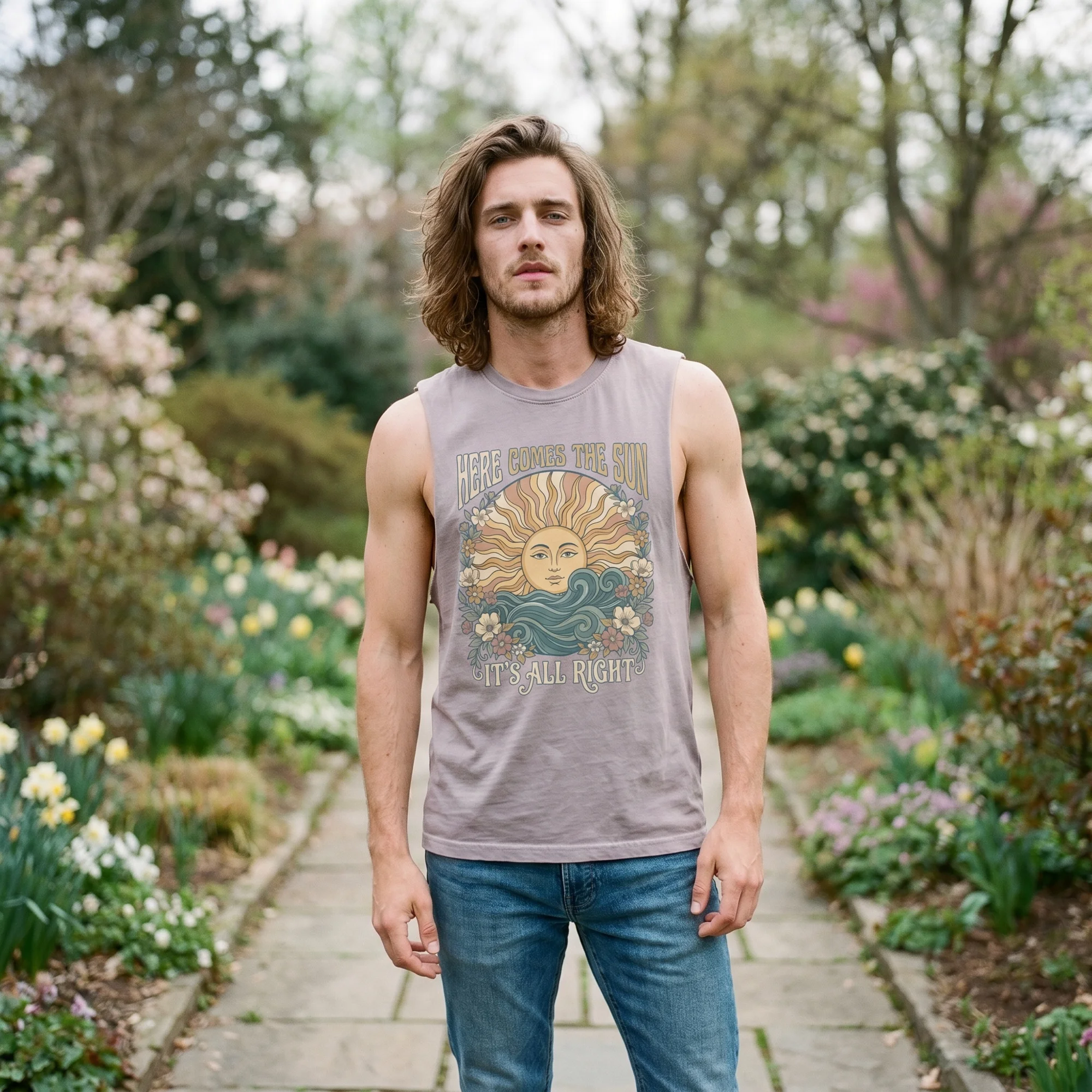 Man with long wavy hair wearing an orchid Here Comes the Sun tank top, front view, standing on a garden path lined with spring flowers
