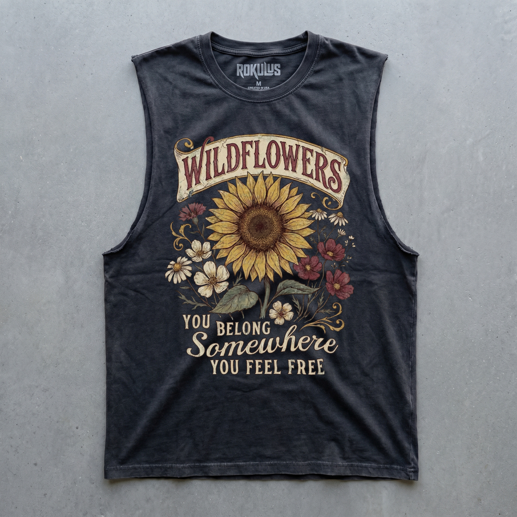 Black cut-off muscle tank laid flat on concrete, showing the Wildflowers sunflower graphic with prairie blooms and "You Belong Somewhere You Feel Free" lettering, plus the Rokulus inner-neck label