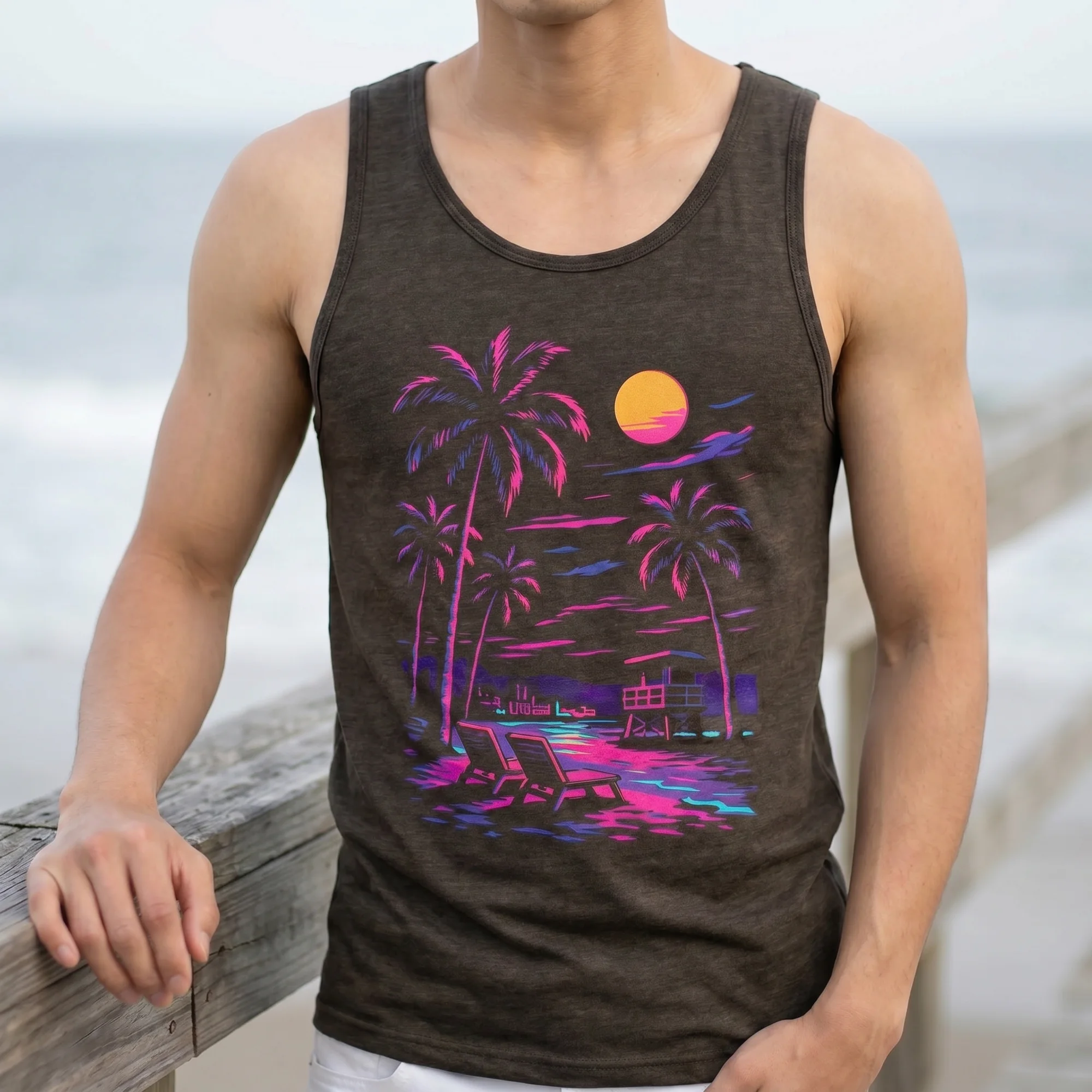 Close-up front view of a man wearing a charcoal black triblend tank top with the Retro Neon Beach palm tree and sunset graphic, beach in the background