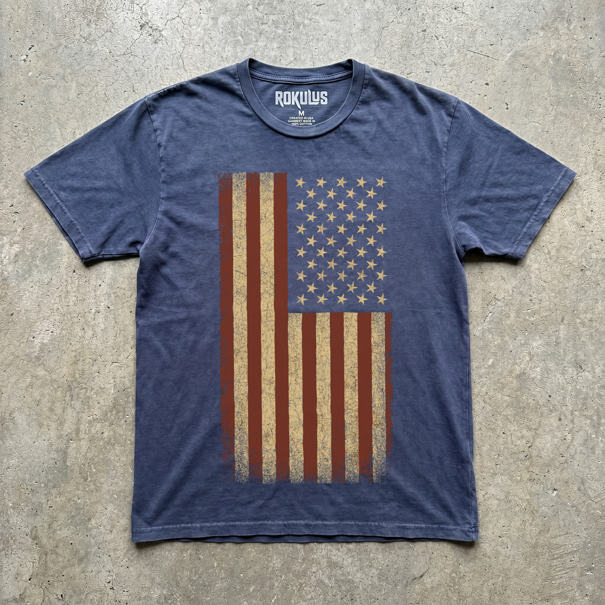 Faded denim blue vintage wash t-shirt laid flat on concrete, showing the distressed American flag graphic in oxblood red and aged cream