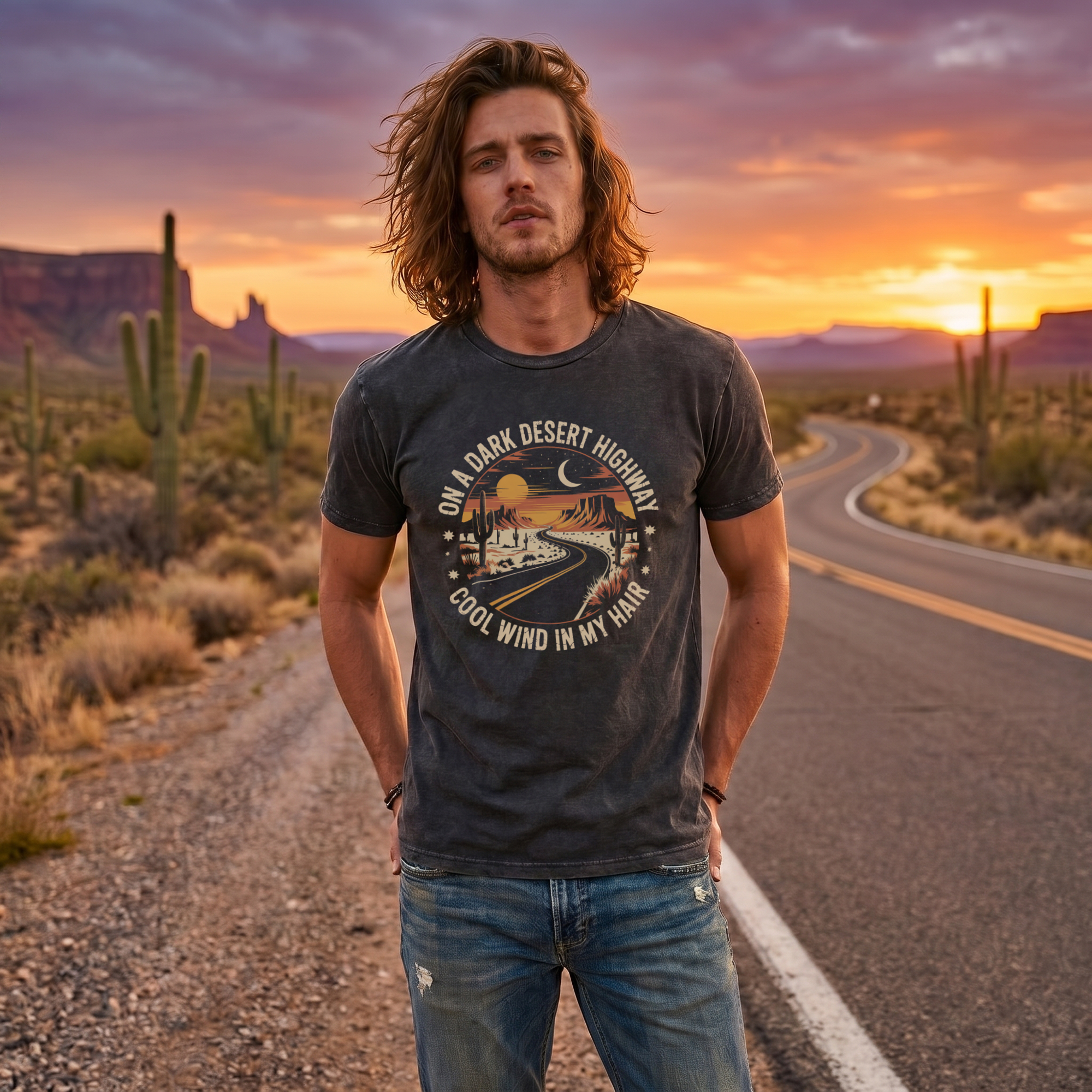 Man wearing a black vintage wash t-shirt with a desert highway graphic, front view, standing on a desert road at sunset with cacti and mesas