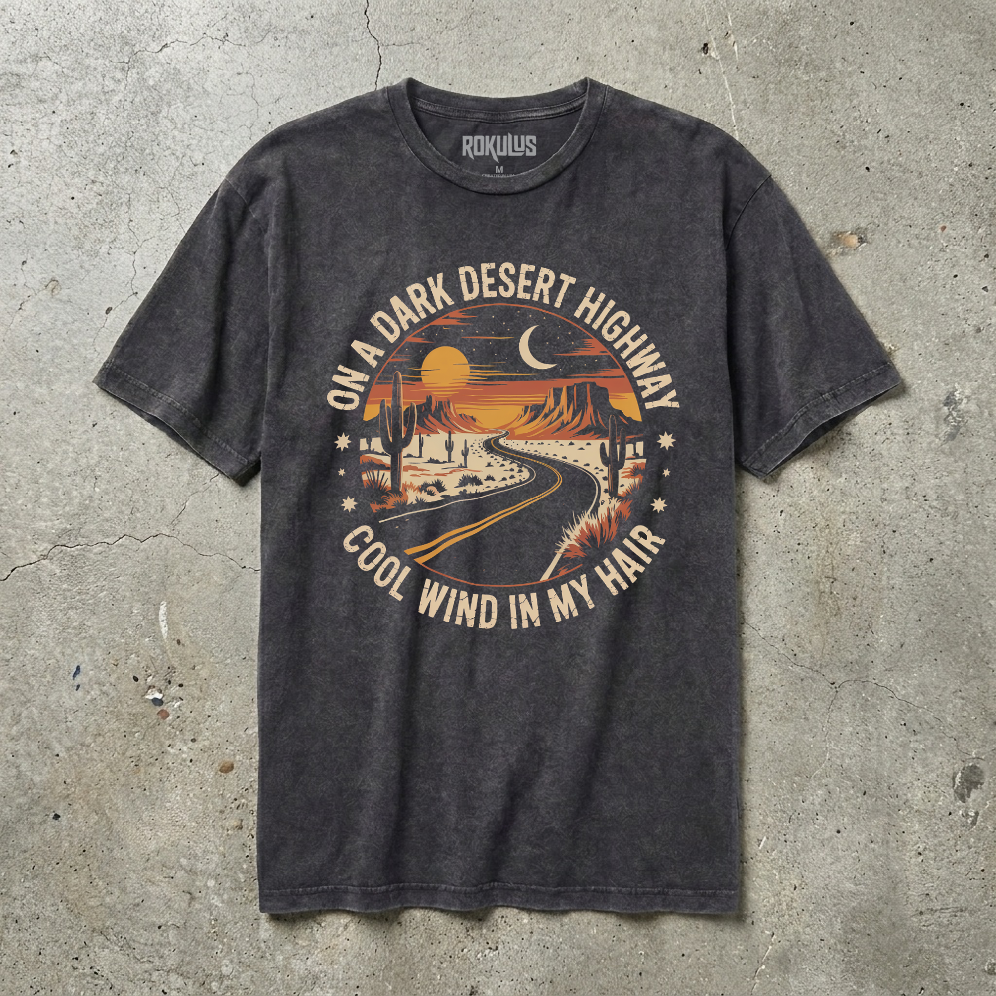 Black vintage wash t-shirt laid flat on concrete, showing a circular desert landscape graphic with sunset, moon, mesas, and saguaro cacti
