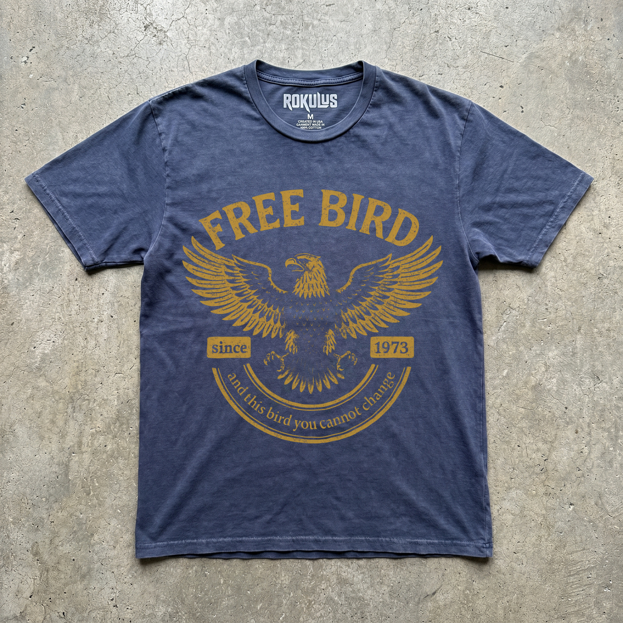 Faded denim blue vintage wash t-shirt laid flat on concrete, showing the gold soaring eagle graphic and vintage banner typography
