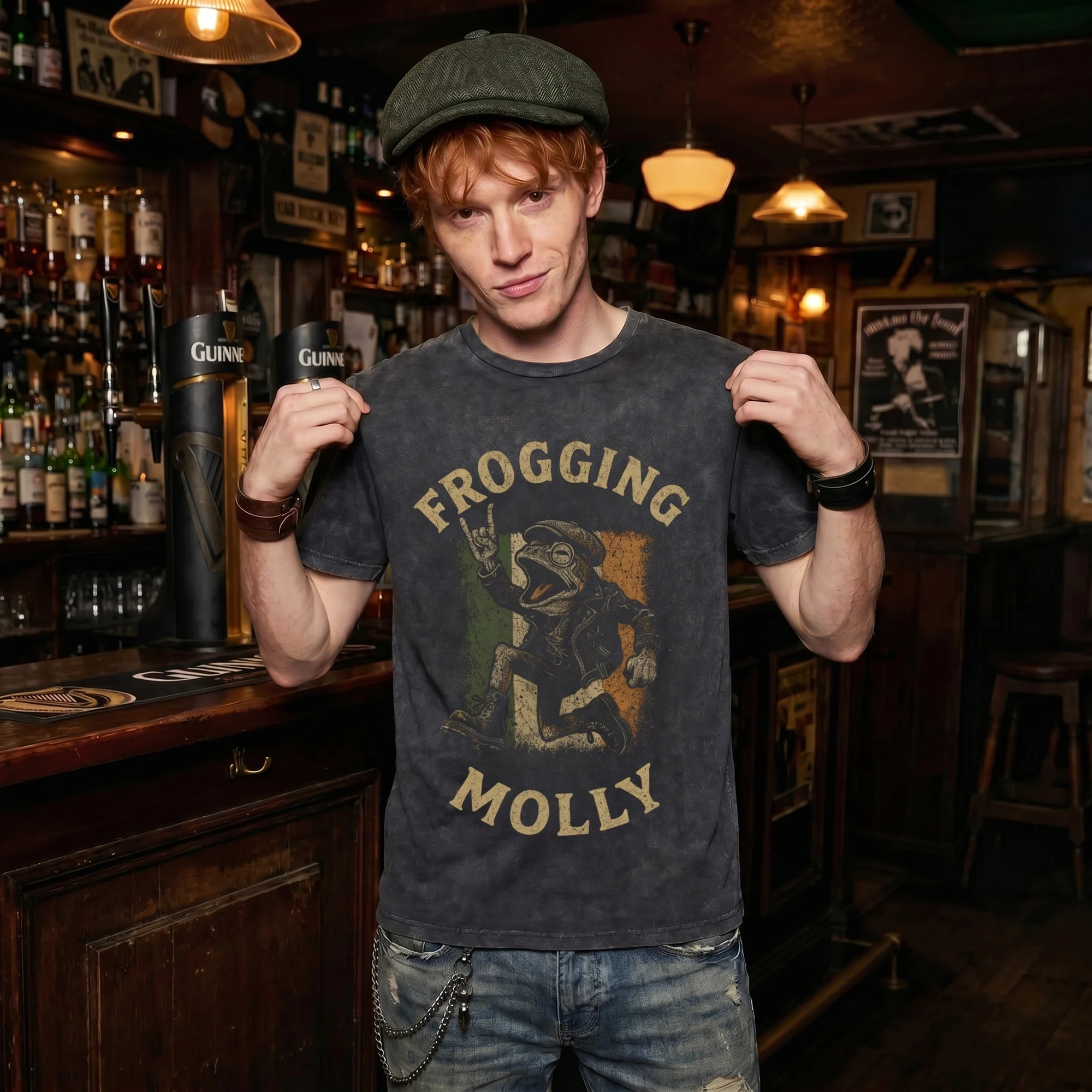 Man in a green flat cap wearing a black vintage wash Frogging Molly t-shirt, front view, holding the tee open in a wood-paneled Irish pub with Guinness taps