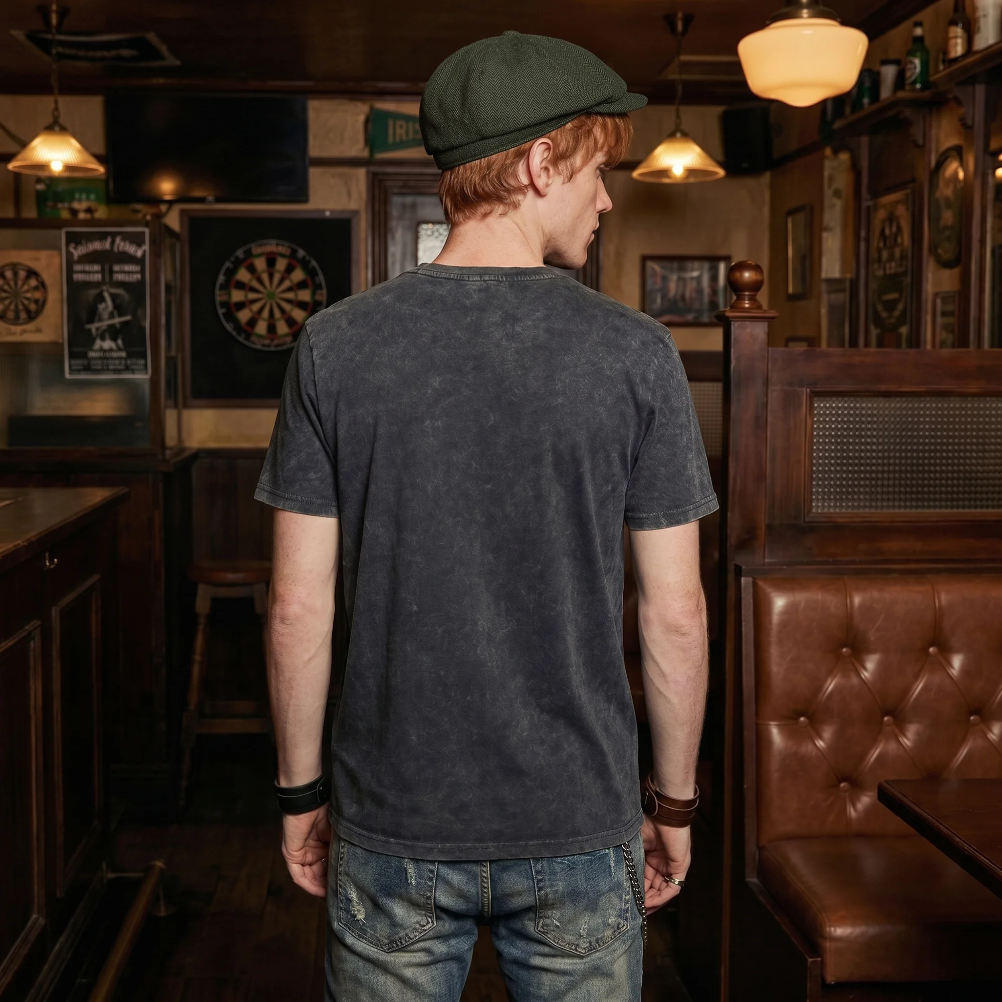 Back view of a man in a green flat cap wearing a black vintage wash t-shirt, standing in an Irish pub with a dartboard on the wall