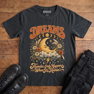 Washed black Rokulus Dreams vintage graphic tee laid flat on wood with black boots and jeans, front print showing a sleeping crescent moon on a storm cloud with wildflowers and moon phases under “Dreams” lettering and “Thunder Only Happens When It’s Rainin’”