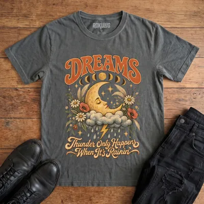 Washed charcoal Rokulus Dreams vintage graphic tee laid flat on wood with black boots and jeans, front print showing a sleeping crescent moon on a storm cloud with wildflowers and moon phases under “Dreams” lettering and “Thunder Only Happens When It’s Rainin’”