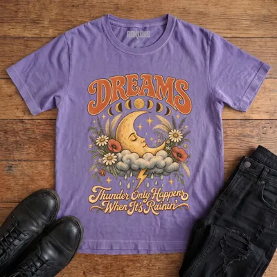 Washed purple Rokulus Dreams vintage graphic tee laid flat on wood with black boots and jeans, front print showing a sleeping crescent moon on a storm cloud with wildflowers and moon phases under “Dreams” lettering and “Thunder Only Happens When It’s Rainin’”
