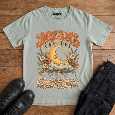Washed thyme Rokulus Dreams vintage graphic tee laid flat on wood with black boots and jeans, front print showing a sleeping crescent moon on a storm cloud with wildflowers and moon phases under “Dreams” lettering and “Thunder Only Happens When It’s Rainin’”