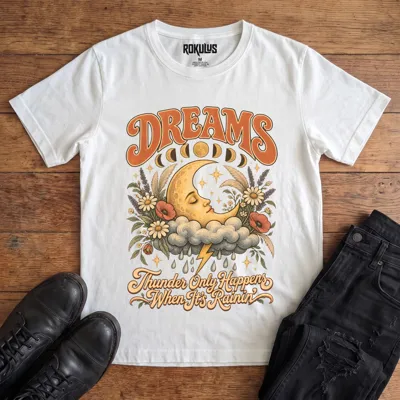 White Rokulus Dreams vintage graphic tee laid flat on wood with black boots and jeans, front print showing a sleeping crescent moon on a storm cloud with wildflowers and moon phases under “Dreams” lettering and “Thunder Only Happens When It’s Rainin’”