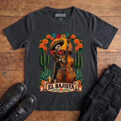 Washed black Rokulus El Bajista vintage graphic tee laid flat on wood with black boots and jeans, front print showing a Day of the Dead skeleton mariachi in a sombrero playing an upright bass with marigolds, cactus, and papel picado above an “El Bajista” banner