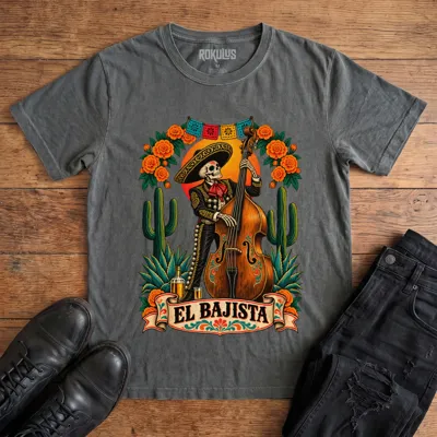 Washed charcoal Rokulus El Bajista vintage graphic tee laid flat on wood with black boots and jeans, front print showing a Day of the Dead skeleton mariachi in a sombrero playing an upright bass with marigolds, cactus, and papel picado above an “El Bajista” banner