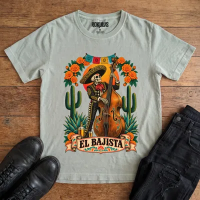 Washed thyme Rokulus El Bajista vintage graphic tee laid flat on wood with black boots and jeans, front print showing a Day of the Dead skeleton mariachi in a sombrero playing an upright bass with marigolds, cactus, and papel picado above an “El Bajista” banner