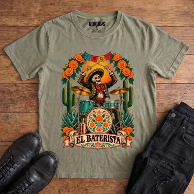 Washed olive Rokulus El Baterista vintage graphic tee laid flat on wood with black boots and jeans, front print showing a Day of the Dead skeleton mariachi in a sombrero playing drums with marigolds, cactus, and papel picado above an “El Baterista” banner