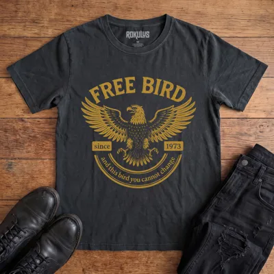 Washed black Rokulus Free Bird vintage graphic tee laid flat on wood with black boots and jeans, front print showing a spread-winged eagle in distressed gold with arched "Free Bird" lettering and vintage banner type