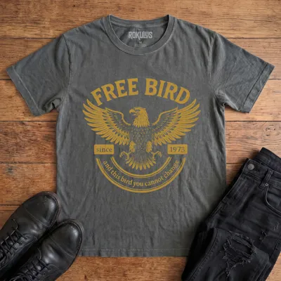 Washed charcoal Rokulus Free Bird vintage graphic tee laid flat on wood with black boots and jeans, front print showing a spread-winged eagle in distressed gold with arched "Free Bird" lettering and vintage banner type