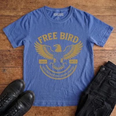 Washed royal blue Rokulus Free Bird vintage graphic tee laid flat on wood with black boots and jeans, front print showing a spread-winged eagle in distressed gold with arched "Free Bird" lettering and vintage banner type