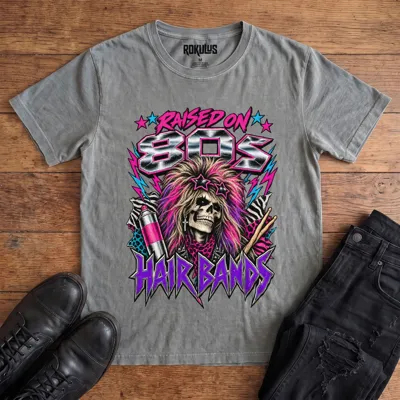 Washed grey Rokulus Raised On 80s Hair Bands vintage graphic tee laid flat on wood with black boots and jeans, front print showing a big-haired skeleton glam rocker in star sunglasses with hairspray, drumsticks, and animal print in neon and chrome