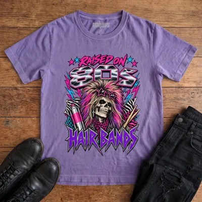 Washed purple Rokulus Raised On 80s Hair Bands vintage graphic tee laid flat on wood with black boots and jeans, front print showing a big-haired skeleton glam rocker in star sunglasses with hairspray, drumsticks, and animal print in neon and chrome