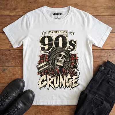 White Rokulus Raised On 90s Grunge vintage graphic tee laid flat on wood with black boots and jeans, front print showing a long-haired skull in a beanie and headphones on red plaid flannel with a cassette tape under “Raised On 90s Grunge” lettering