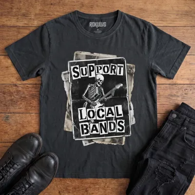 Washed black Rokulus Support Local Bands vintage graphic tee laid flat on wood with black boots and jeans, front print showing a torn punk flyer with a skeleton playing electric guitar and "Support Local Bands" in ransom-note lettering