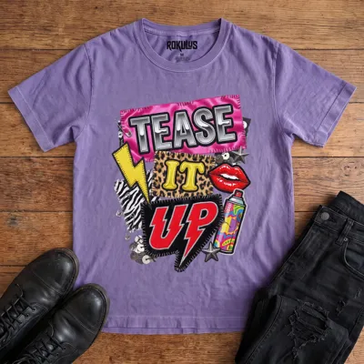 Washed purple Rokulus Tease It Up vintage graphic tee laid flat on wood with black boots and jeans, front print showing an 80s glam collage with leopard and zebra patches, red lips, hairspray, and lightning bolts under chrome “Tease It Up” lettering