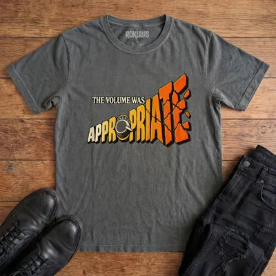 Washed charcoal Rokulus The Volume Was Appropriate vintage graphic tee laid flat on wood with black boots and jeans, front print reading “The Volume Was Appropriate” with a cranked amp knob and shattering orange-to-yellow lettering