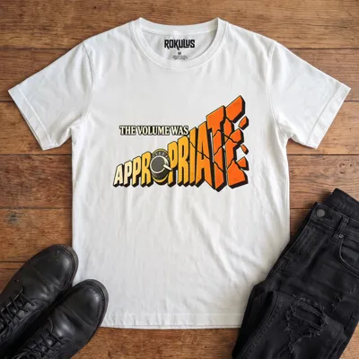 White Rokulus The Volume Was Appropriate vintage graphic tee laid flat on wood with black boots and jeans, front print reading “The Volume Was Appropriate” with a cranked amp knob and shattering orange-to-yellow lettering