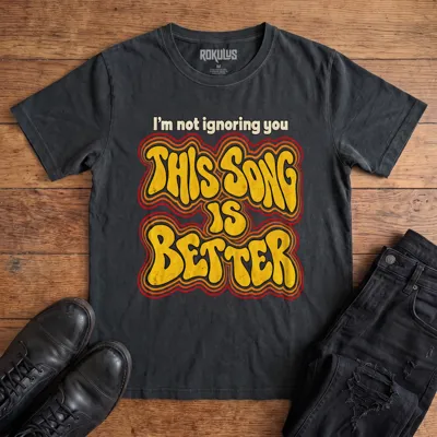 Washed black Rokulus This Song Is Better vintage graphic tee laid flat on wood with black boots and jeans, front print reading “I’m not ignoring you, this song is better” in groovy 70s bubble lettering with a rainbow outline