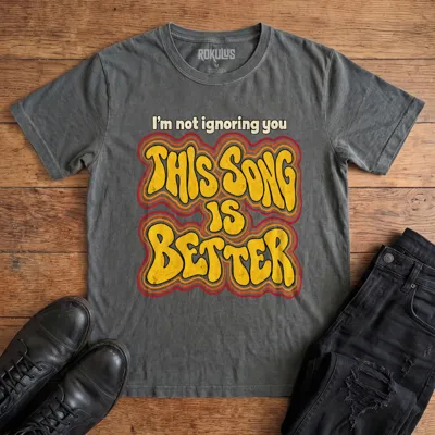 Washed charcoal Rokulus This Song Is Better vintage graphic tee laid flat on wood with black boots and jeans, front print reading “I’m not ignoring you, this song is better” in groovy 70s bubble lettering with a rainbow outline