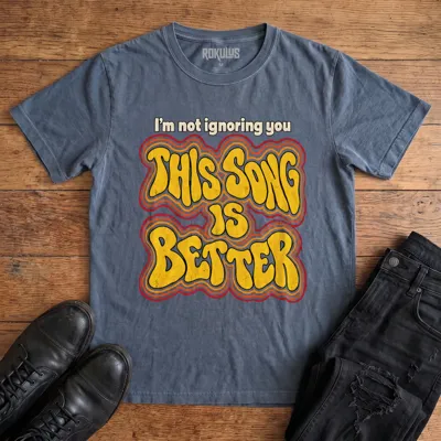 Washed navy blue Rokulus This Song Is Better vintage graphic tee laid flat on wood with black boots and jeans, front print reading “I’m not ignoring you, this song is better” in groovy 70s bubble lettering with a rainbow outline