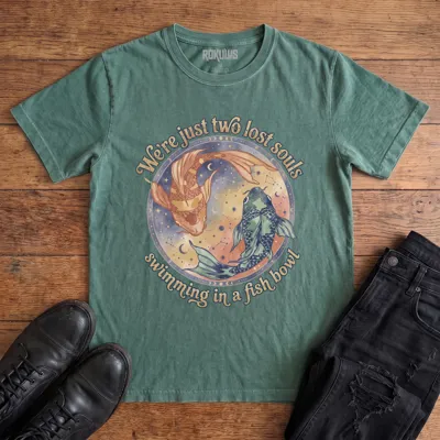 Washed pine Rokulus Two Lost Souls vintage graphic tee laid flat on wood with black boots and jeans, front print showing two watercolor koi in a celestial ring of moon phases and constellations with "We're just two lost souls swimming in a fish bowl" in retro script