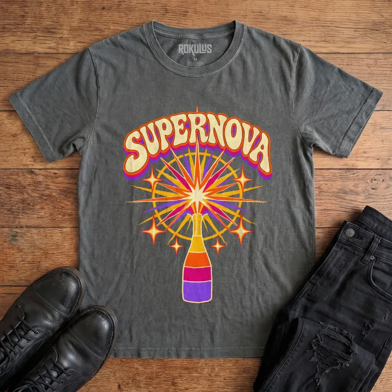 Washed charcoal Rokulus Champagne Supernova vintage graphic tee laid flat on wood with black boots and jeans, front print showing a champagne bottle erupting in a psychedelic sunburst of purple, pink, and orange under groovy “Supernova” lettering
