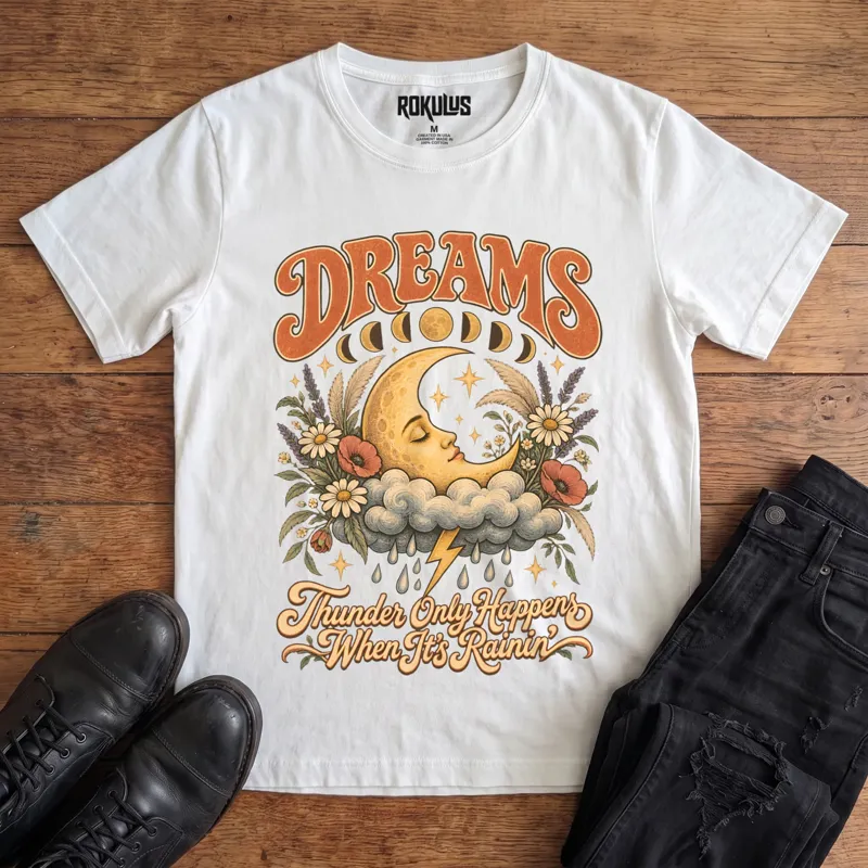 White Rokulus Dreams vintage graphic tee laid flat on wood with black boots and jeans, front print showing a sleeping crescent moon on a storm cloud with wildflowers and moon phases under “Dreams” lettering and “Thunder Only Happens When It’s Rainin’”
