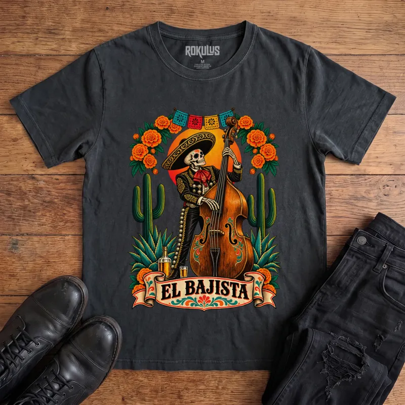 Washed black Rokulus El Bajista vintage graphic tee laid flat on wood with black boots and jeans, front print showing a Day of the Dead skeleton mariachi in a sombrero playing an upright bass with marigolds, cactus, and papel picado above an “El Bajista” banner
