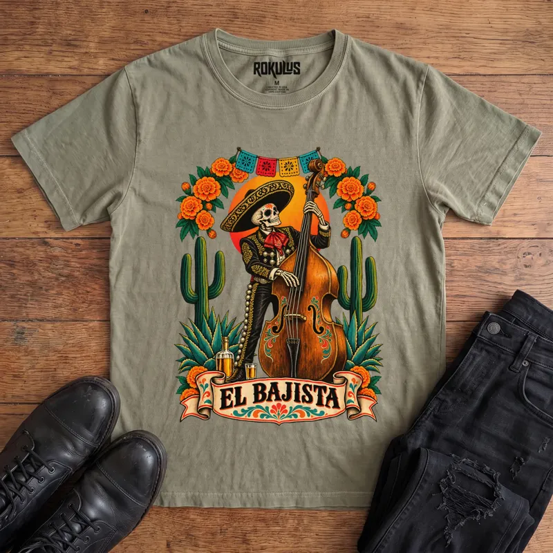 Washed olive Rokulus El Bajista vintage graphic tee laid flat on wood with black boots and jeans, front print showing a Day of the Dead skeleton mariachi in a sombrero playing an upright bass with marigolds, cactus, and papel picado above an “El Bajista” banner