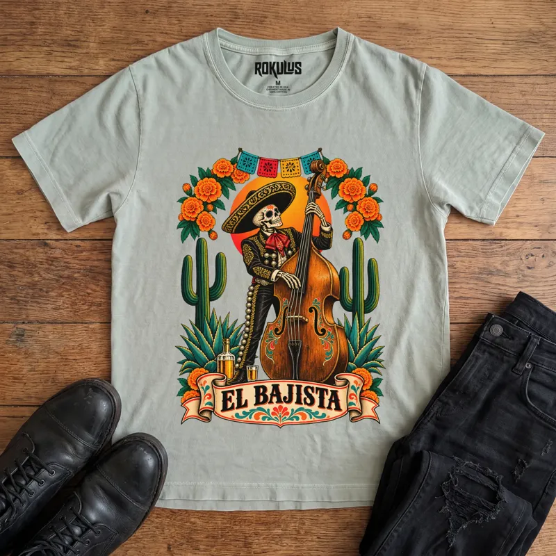 Washed thyme Rokulus El Bajista vintage graphic tee laid flat on wood with black boots and jeans, front print showing a Day of the Dead skeleton mariachi in a sombrero playing an upright bass with marigolds, cactus, and papel picado above an “El Bajista” banner