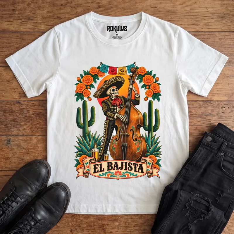 White Rokulus El Bajista vintage graphic tee laid flat on wood with black boots and jeans, front print showing a Day of the Dead skeleton mariachi in a sombrero playing an upright bass with marigolds, cactus, and papel picado above an “El Bajista” banner