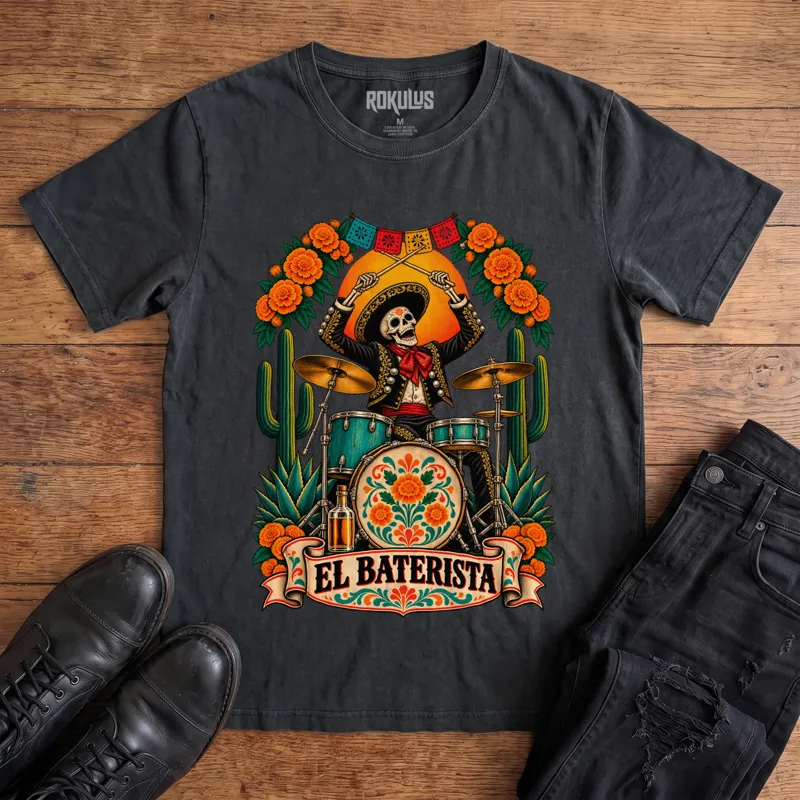Washed black Rokulus El Baterista vintage graphic tee laid flat on wood with black boots and jeans, front print showing a Day of the Dead skeleton mariachi in a sombrero playing drums with marigolds, cactus, and papel picado above an “El Baterista” banner