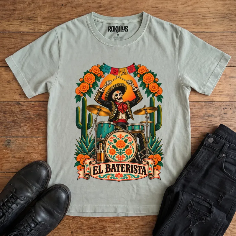 Washed thyme Rokulus El Baterista vintage graphic tee laid flat on wood with black boots and jeans, front print showing a Day of the Dead skeleton mariachi in a sombrero playing drums with marigolds, cactus, and papel picado above an “El Baterista” banner