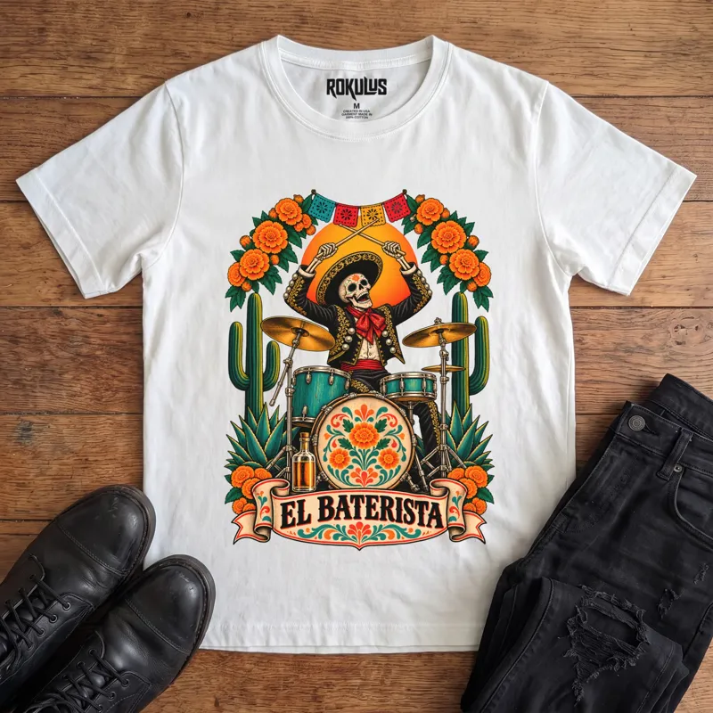 White Rokulus El Baterista vintage graphic tee laid flat on wood with black boots and jeans, front print showing a Day of the Dead skeleton mariachi in a sombrero playing drums with marigolds, cactus, and papel picado above an “El Baterista” banner