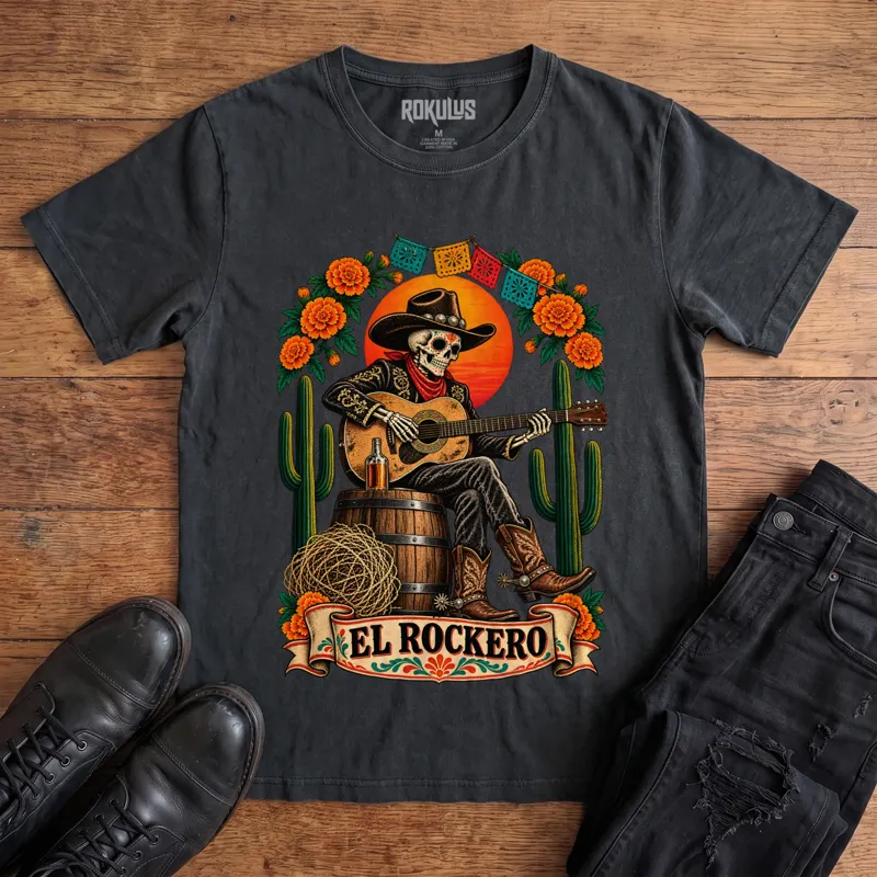 Washed black Rokulus El Rockero vintage graphic tee laid flat on wood with black boots and jeans, front print showing a Day of the Dead skeleton charro in a cowboy hat playing guitar on a barrel with marigolds, cactus, and papel picado above an “El Rockero” banner