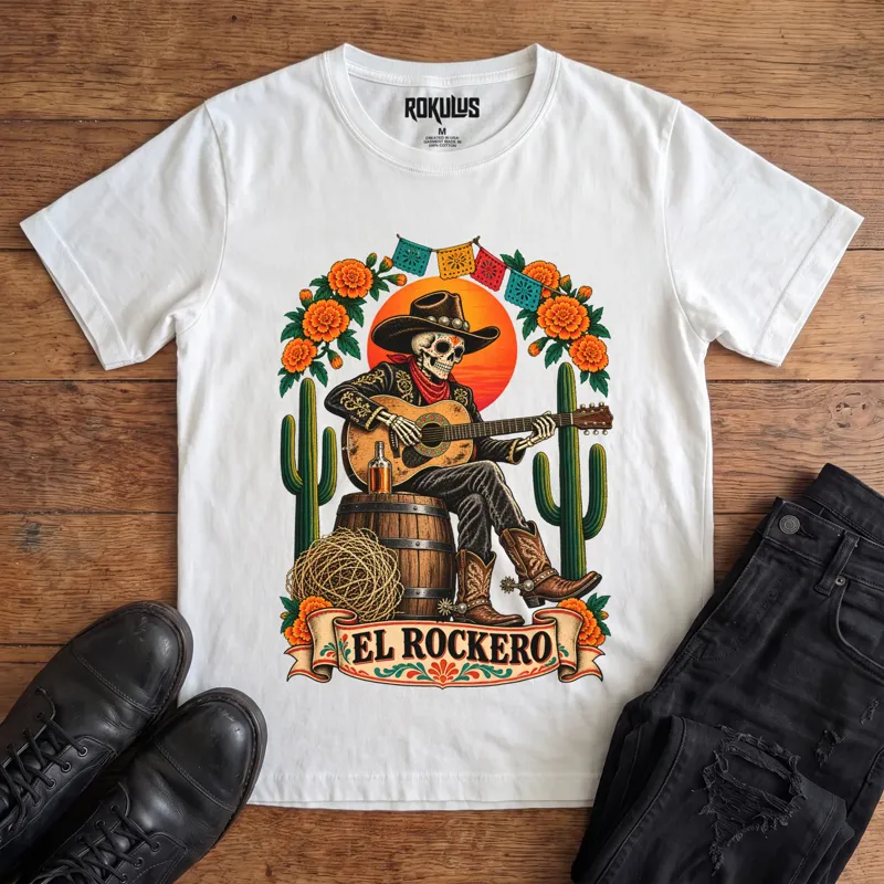 White Rokulus El Rockero vintage graphic tee laid flat on wood with black boots and jeans, front print showing a Day of the Dead skeleton charro in a cowboy hat playing guitar on a barrel with marigolds, cactus, and papel picado above an “El Rockero” banner