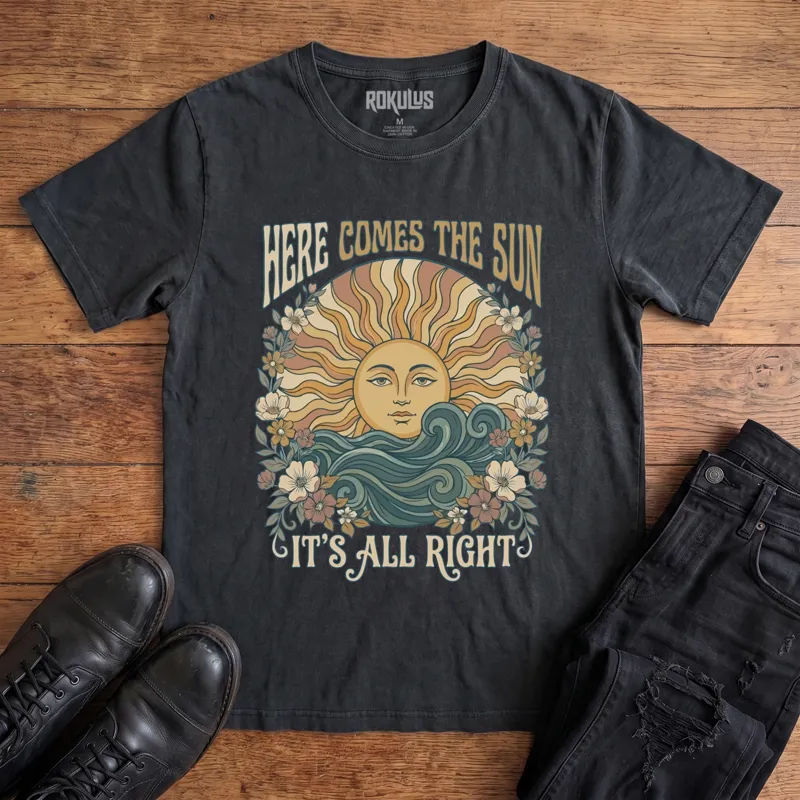 Washed black Rokulus Here Comes The Sun vintage graphic tee laid flat on wood with black boots and jeans, front print showing a boho celestial sun face over ocean waves framed by wildflowers with groovy "Here Comes The Sun It's All Right" lettering