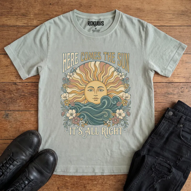 Washed thyme Rokulus Here Comes The Sun vintage graphic tee laid flat on wood with black boots and jeans, front print showing a boho celestial sun face over ocean waves framed by wildflowers with groovy "Here Comes The Sun It's All Right" lettering