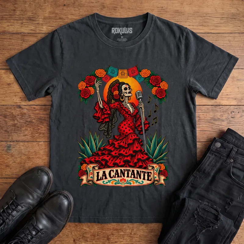 Washed black Rokulus La Cantante vintage graphic tee laid flat on wood with black boots and jeans, front print showing a Day of the Dead skeleton woman in a red flamenco dress singing into a microphone with roses, marigolds, and papel picado above a “La Cantante” banner